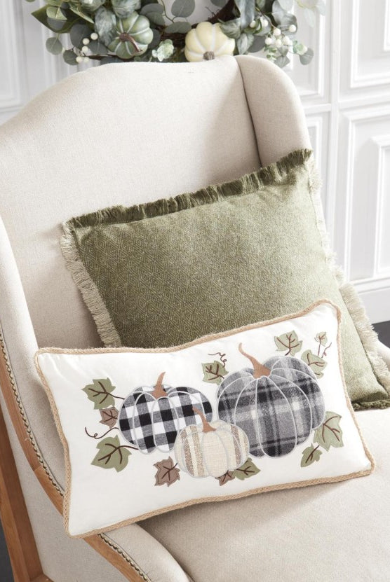 Rectangular cream canvas pillow with plaid pumpkin appliqué and jute trim, displayed on a chair with a green pillow.
