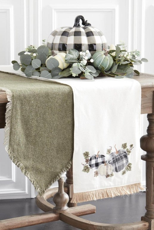 Cream canvas table runner with pumpkin appliqué and jute fringe on a wooden table with a buffalo check pumpkin centerpiece.