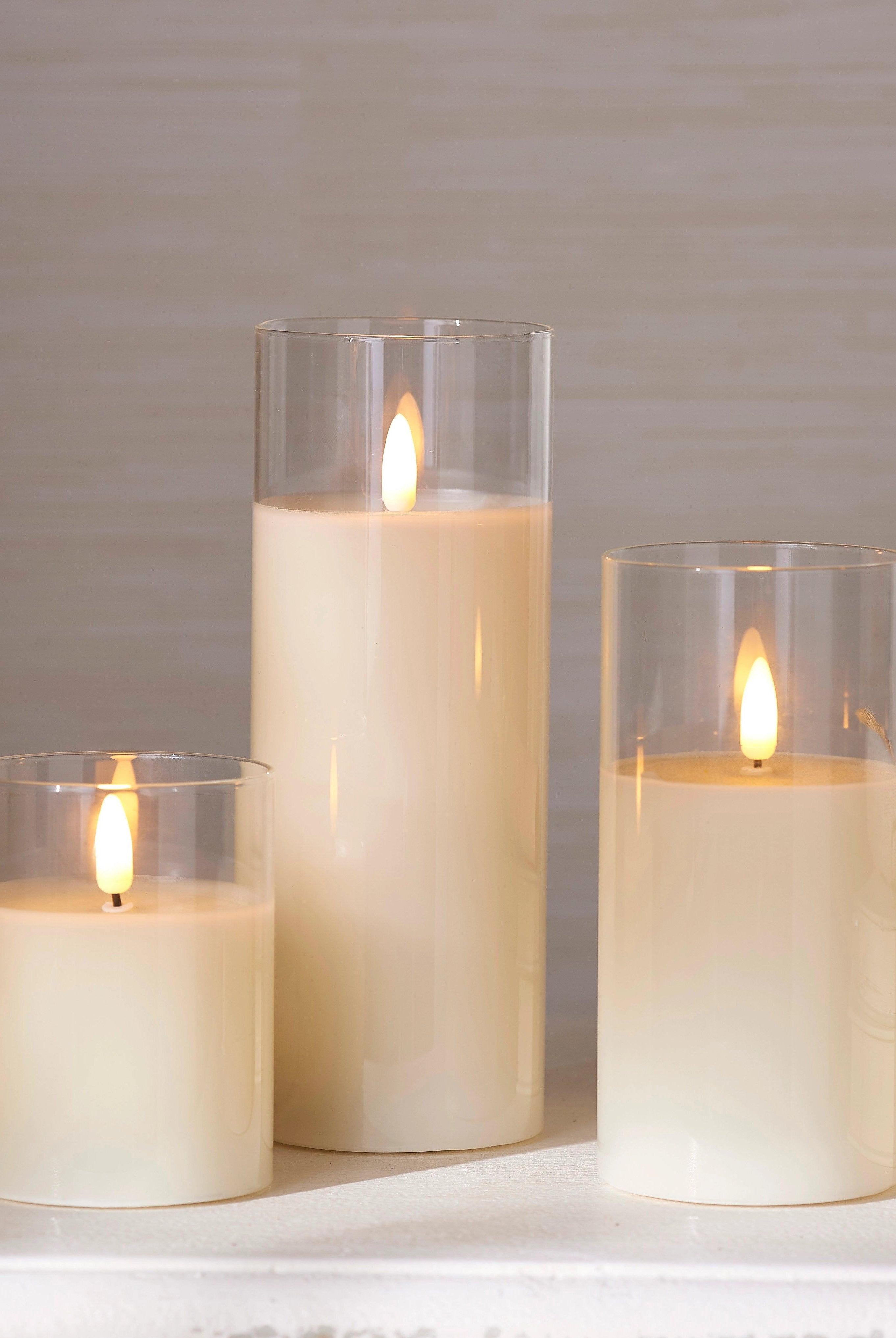 Three ivory LED pillar candles in clear glass holders of varying heights, featuring realistic flickering flames.