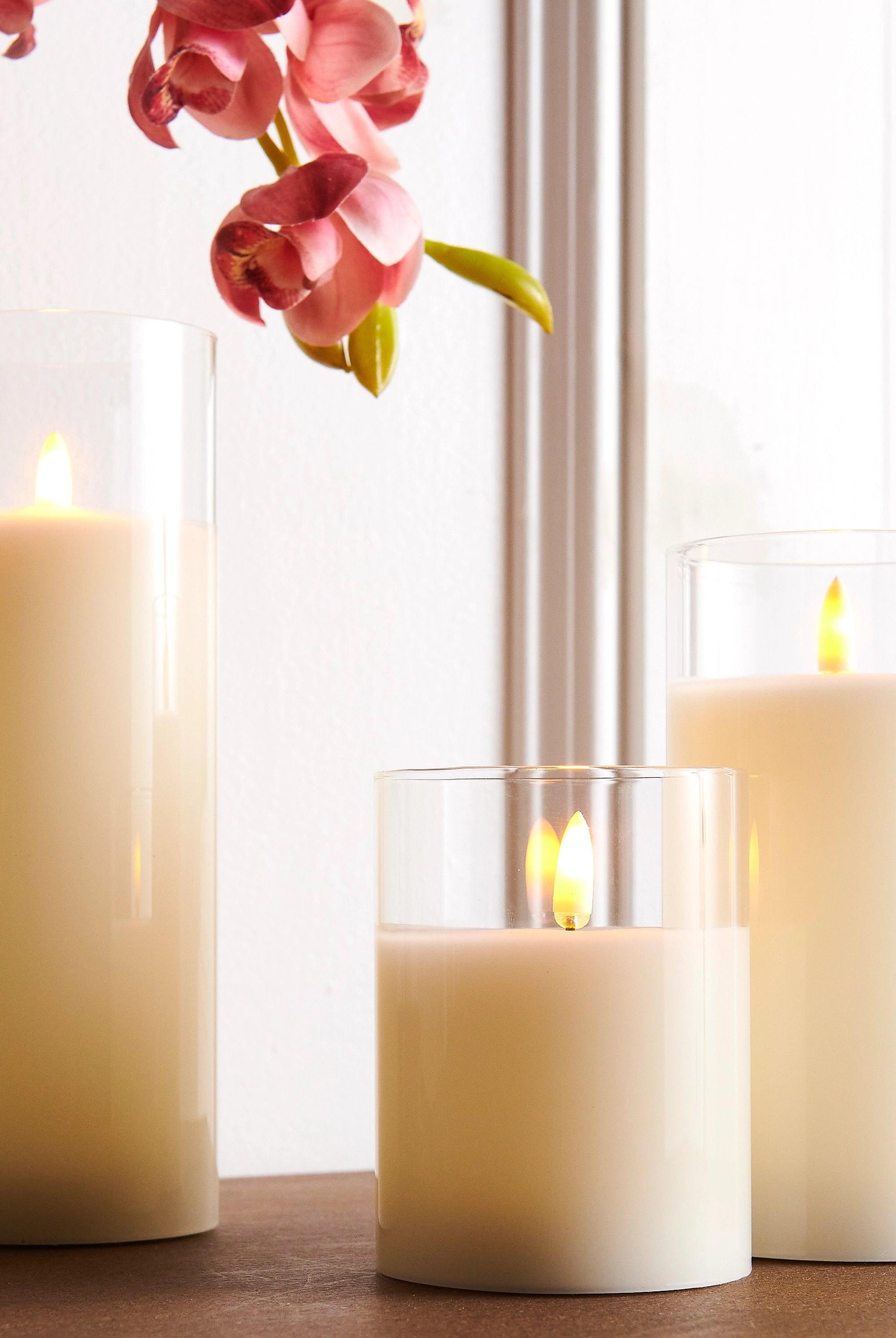 Three ivory LED pillar candles in clear glass holders with realistic flickering flames on a wooden surface.