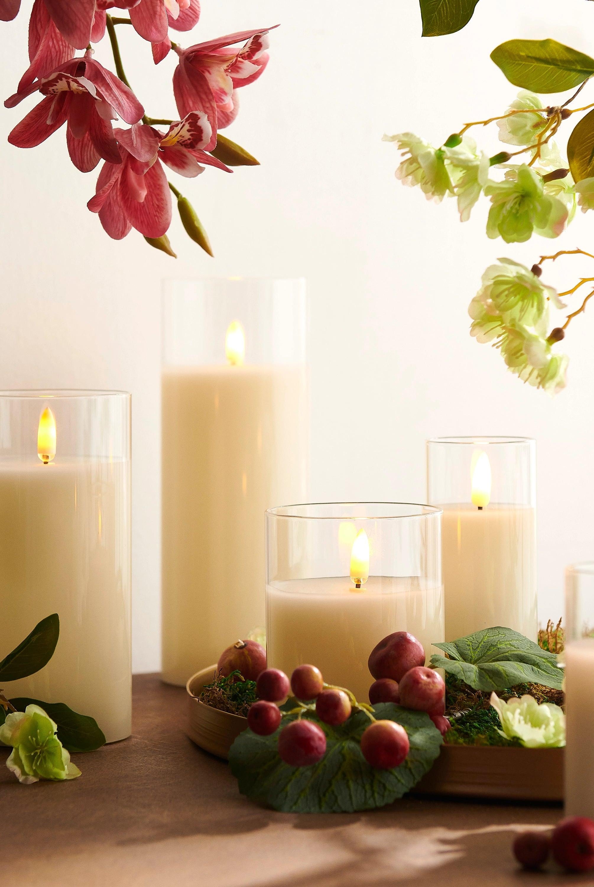 Ivory LED pillar candles in clear glass holders with realistic flickering flames, arranged on a table with floral decor.