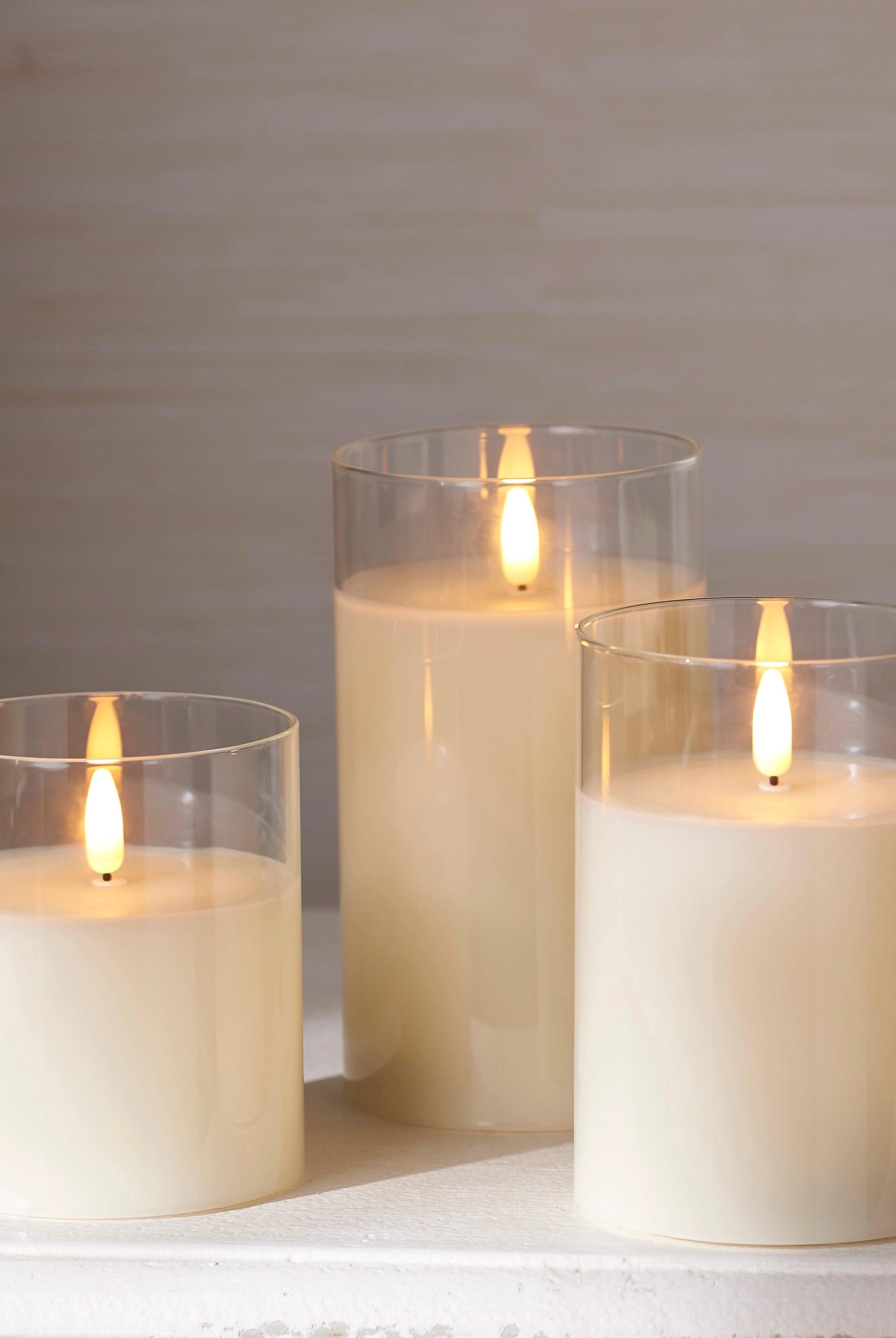 Three ivory LED pillar candles in clear glass holders with realistic flickering flames on a white surface.