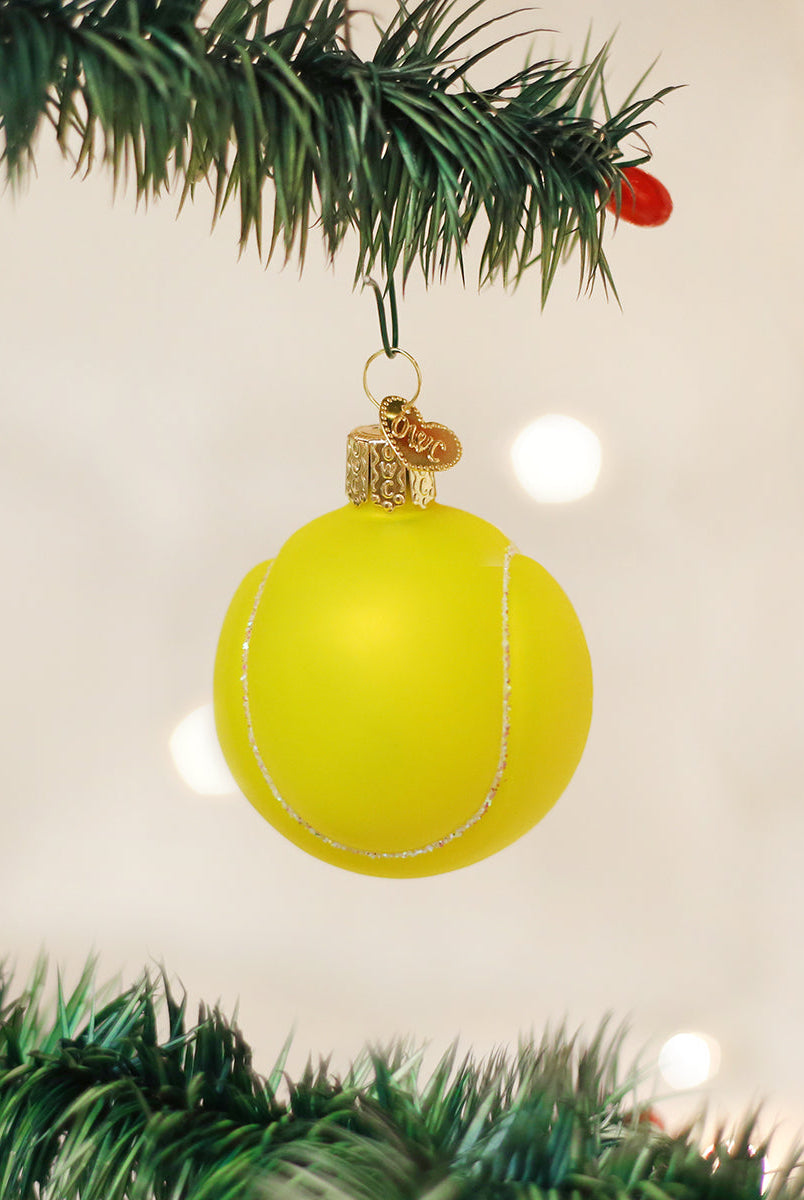 Hand-painted yellow glass tennis ball Christmas ornament with glittery white seams hanging from a pine tree branch.