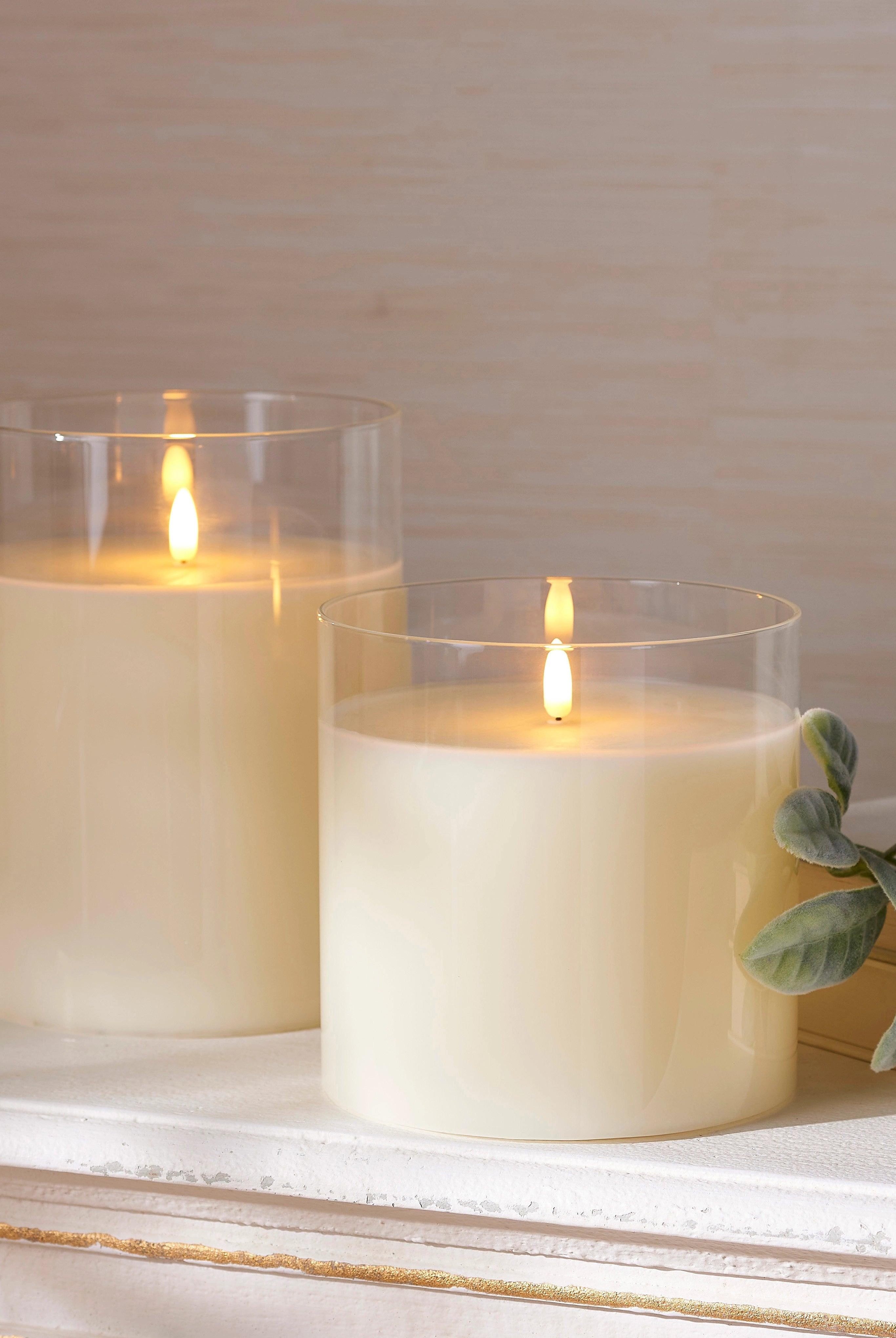 Two 6-inch ivory LED pillar candles in clear glass holders with realistic flickering flames on a white mantel.