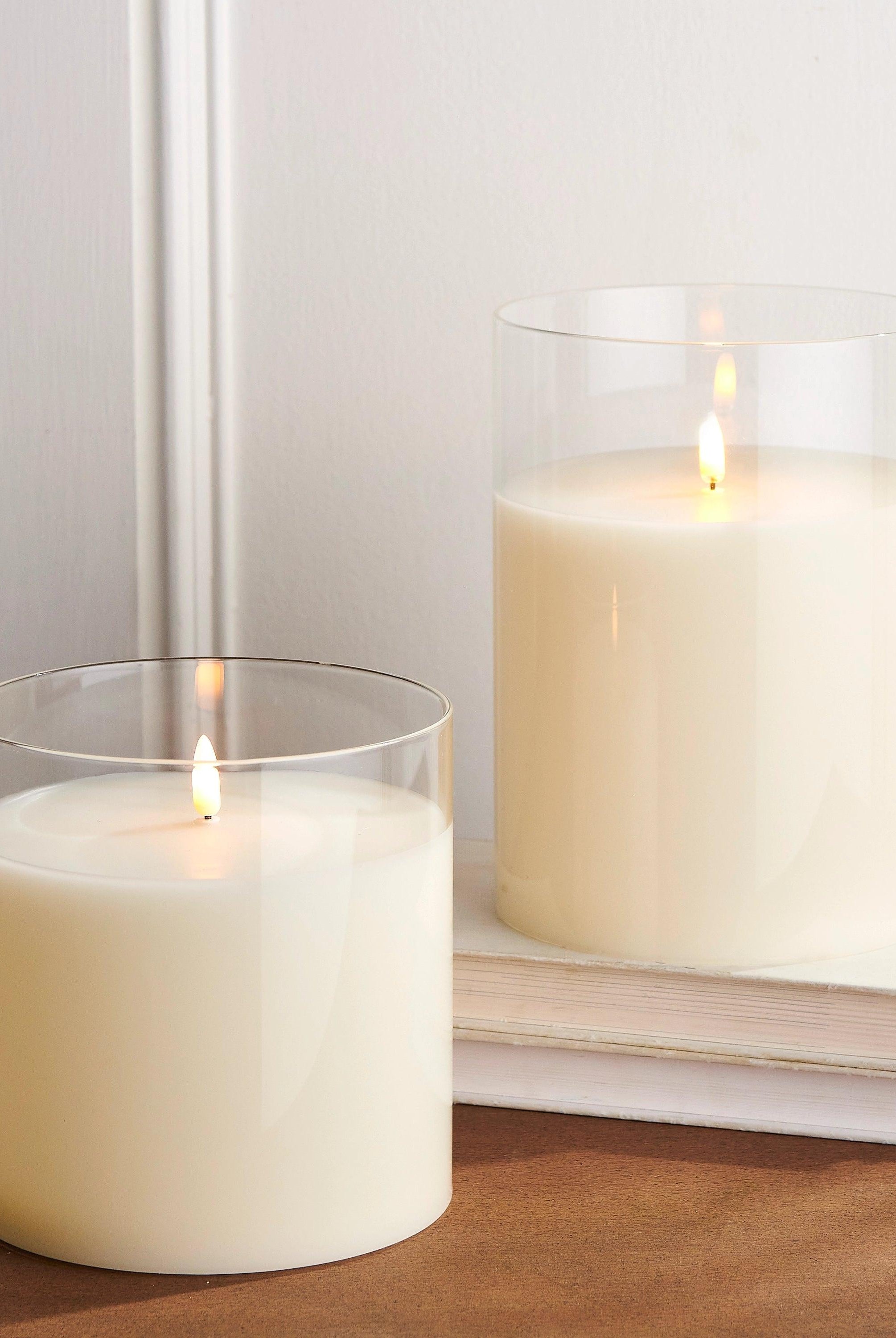 Two ivory LED pillar candles in clear glass holders with realistic flickering flames, set on a wooden surface with books.