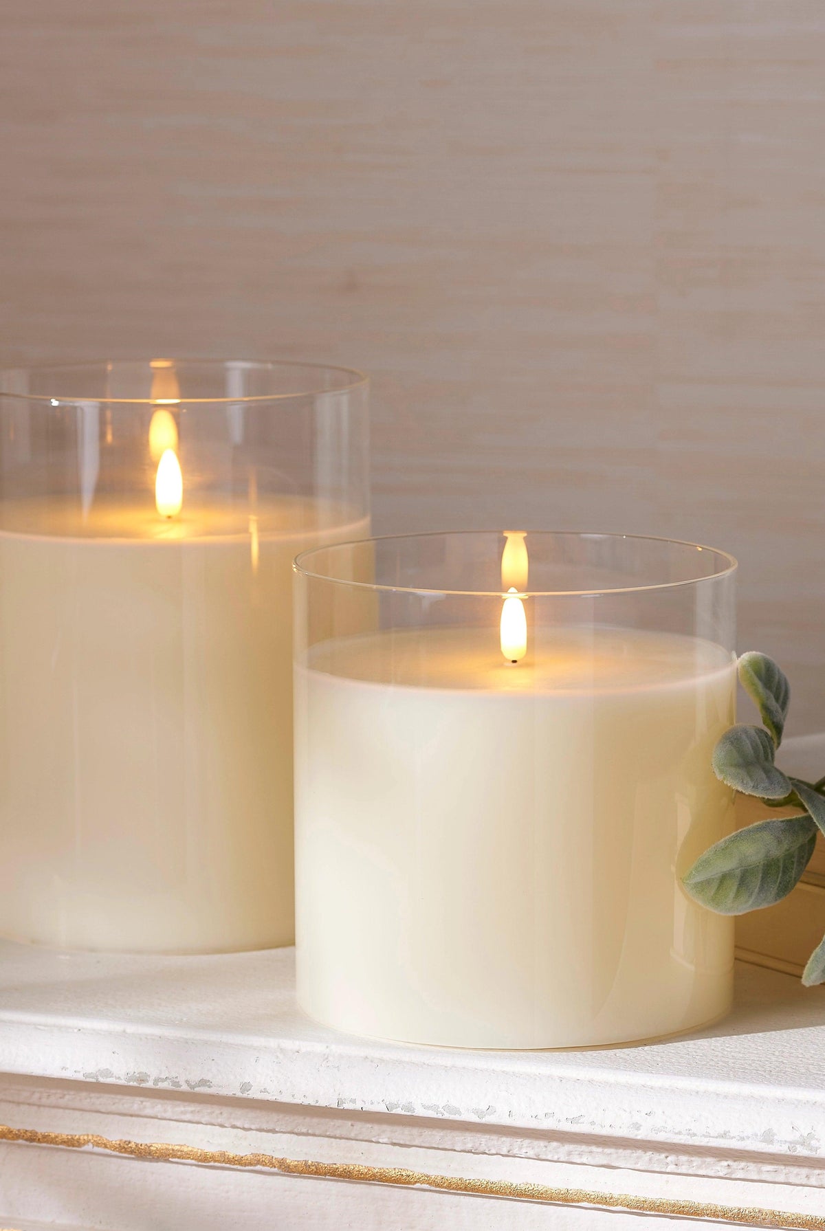 Two ivory LED pillar candles in clear glass holders with flickering flames, set on a white mantel with greenery.