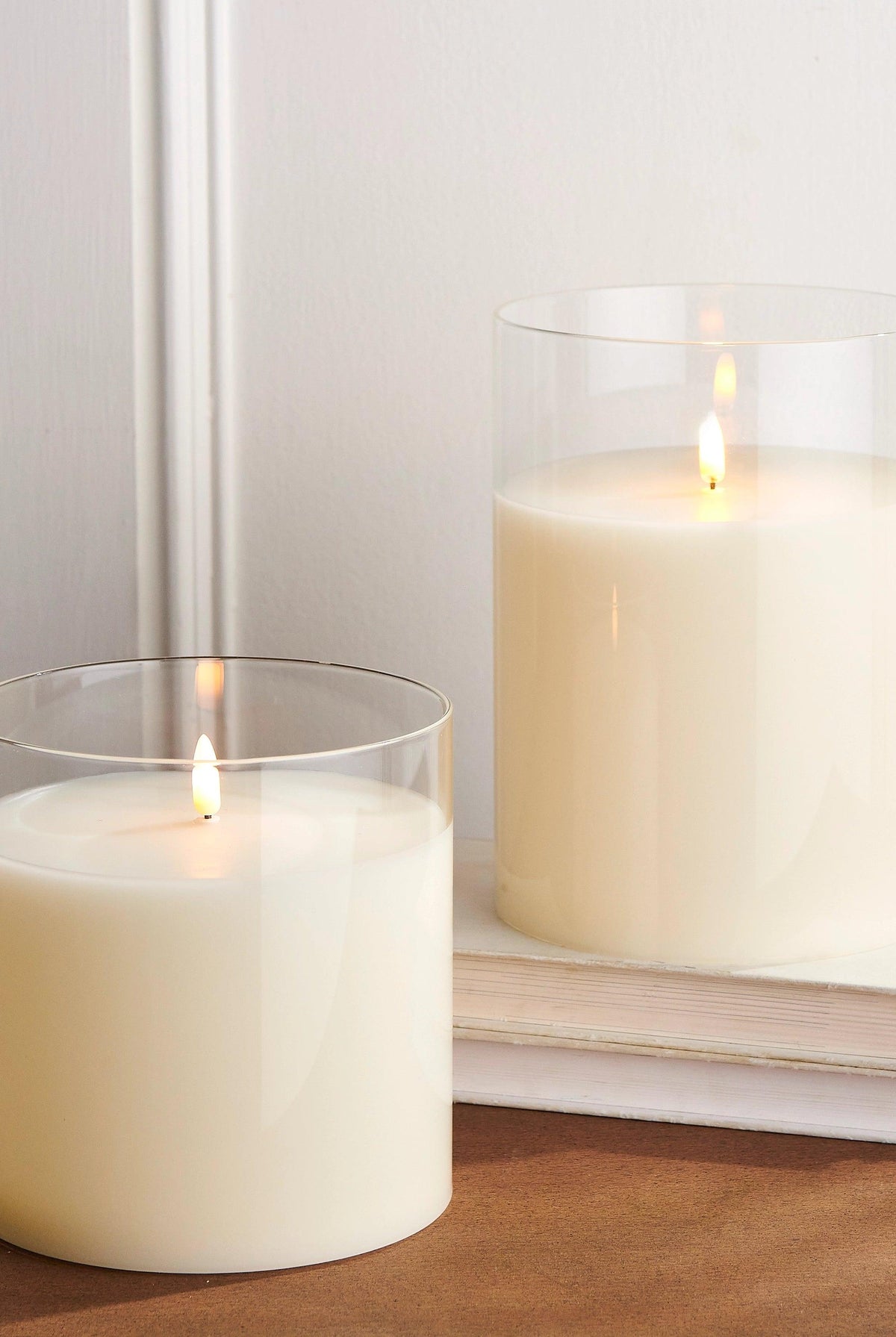 Two ivory LED pillar candles in clear glass holders with realistic flickering flames, set on a wooden surface with books.