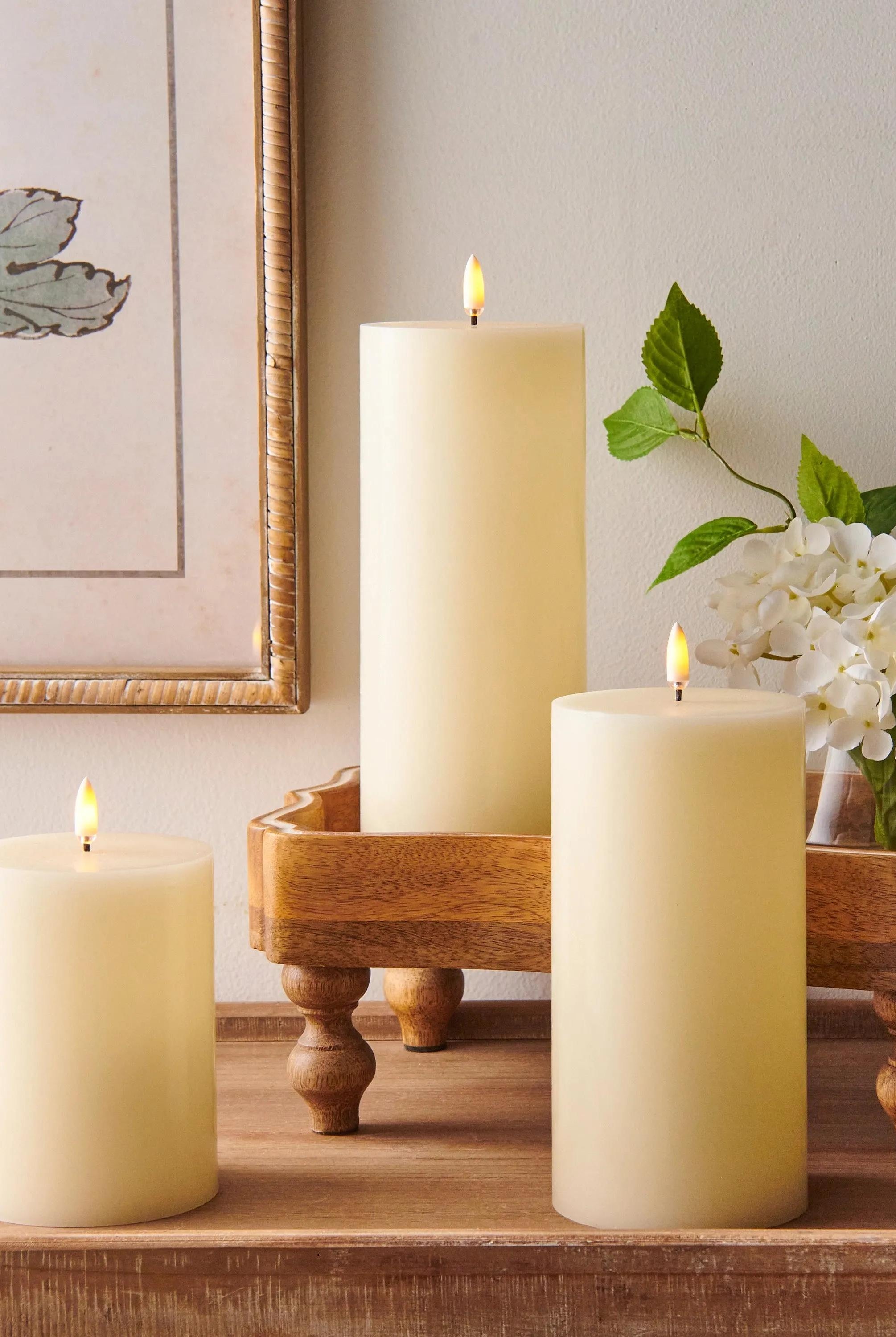 Three ivory wax LED pillar candles with realistic flickering flames on a rustic wooden tray with white flowers.