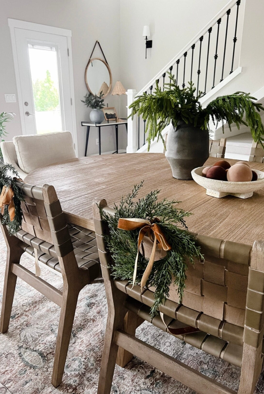 Mini faux cedar wreaths with caramel velvet ribbons hanging on the backs of woven dining chairs in a farmhouse room.