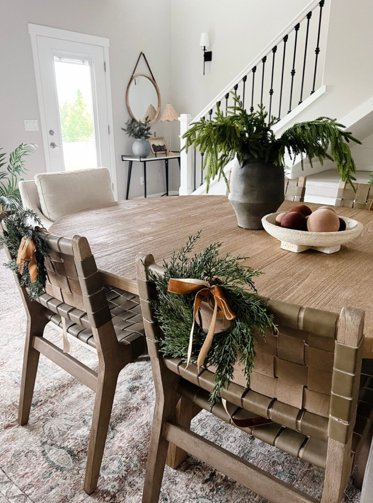 Mini faux cedar wreaths with caramel velvet ribbons hanging on the backs of woven dining chairs in a farmhouse room.