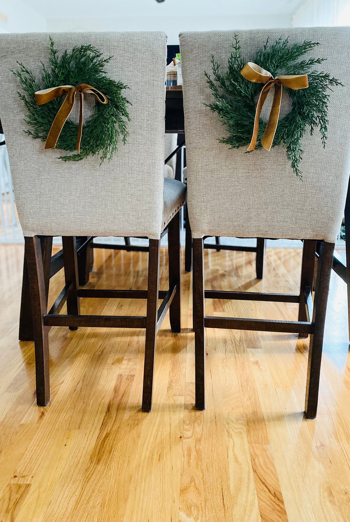 Mini artificial cedar wreaths with elegant gold ribbons hanging on the backs of two upholstered dining chairs.