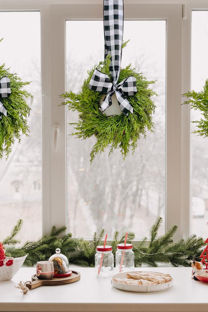 Three mini artificial cedar wreaths with black and white buffalo check ribbons hanging on a window for Christmas.