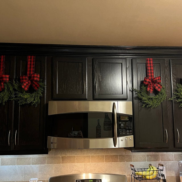 Mini artificial cedar Christmas wreaths with red and black buffalo plaid ribbon hanging on dark kitchen cabinets.