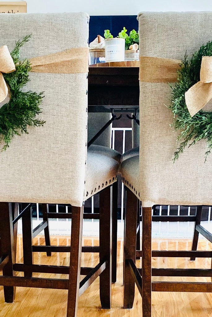 Mini artificial cedar Christmas wreaths with rustic burlap bows tied to the backs of beige upholstered dining chairs.