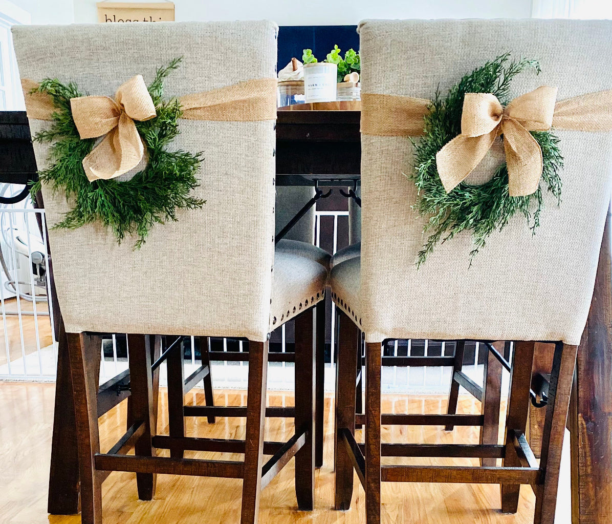 Mini artificial cedar Christmas wreaths with rustic burlap bows tied to the backs of beige upholstered dining chairs.
