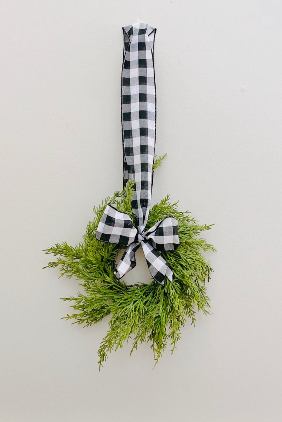 Mini artificial cedar Christmas wreath with a black and white buffalo check ribbon hanging on a plain wall.