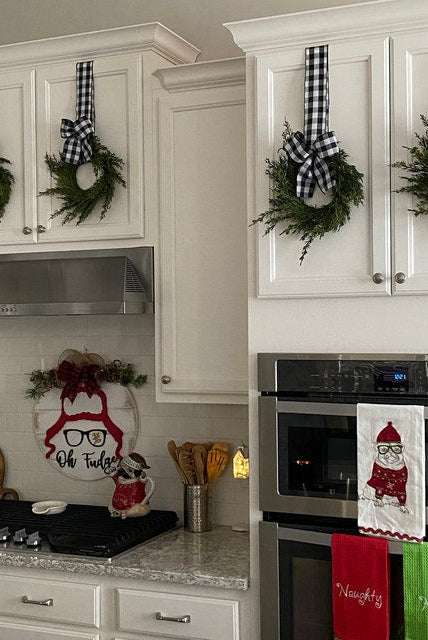 Mini artificial cedar Christmas wreaths with black and white buffalo check ribbons hanging on white kitchen cabinets.