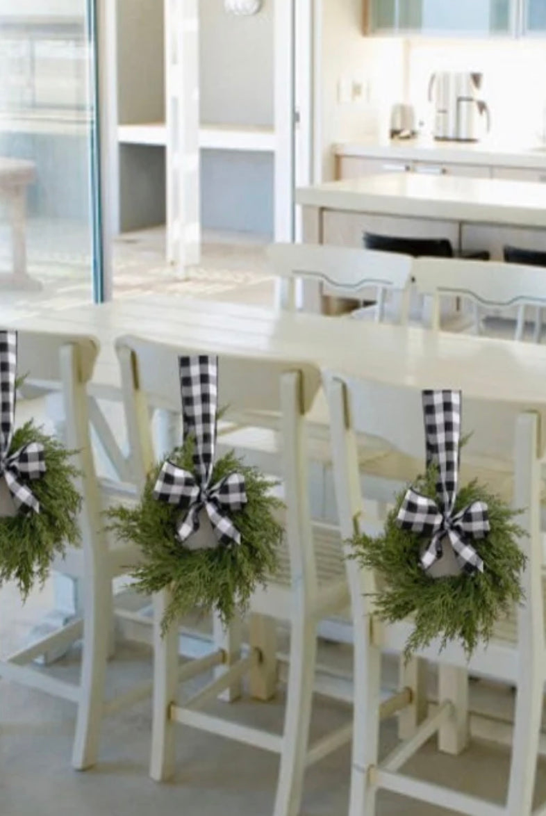 Mini artificial cedar wreaths with black and white buffalo check bows hanging on white dining chairs.