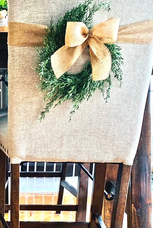 Mini faux cedar Christmas wreath with a rustic burlap bow hanging on a beige upholstered dining chair.