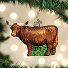 Brown glass Highland cow Christmas ornament with glittery fur texture and long horns hanging on a pine tree.