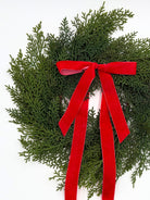 Mini Christmas cabinet wreath made of faux green cedar greenery accented with a long, elegant red velvet ribbon.