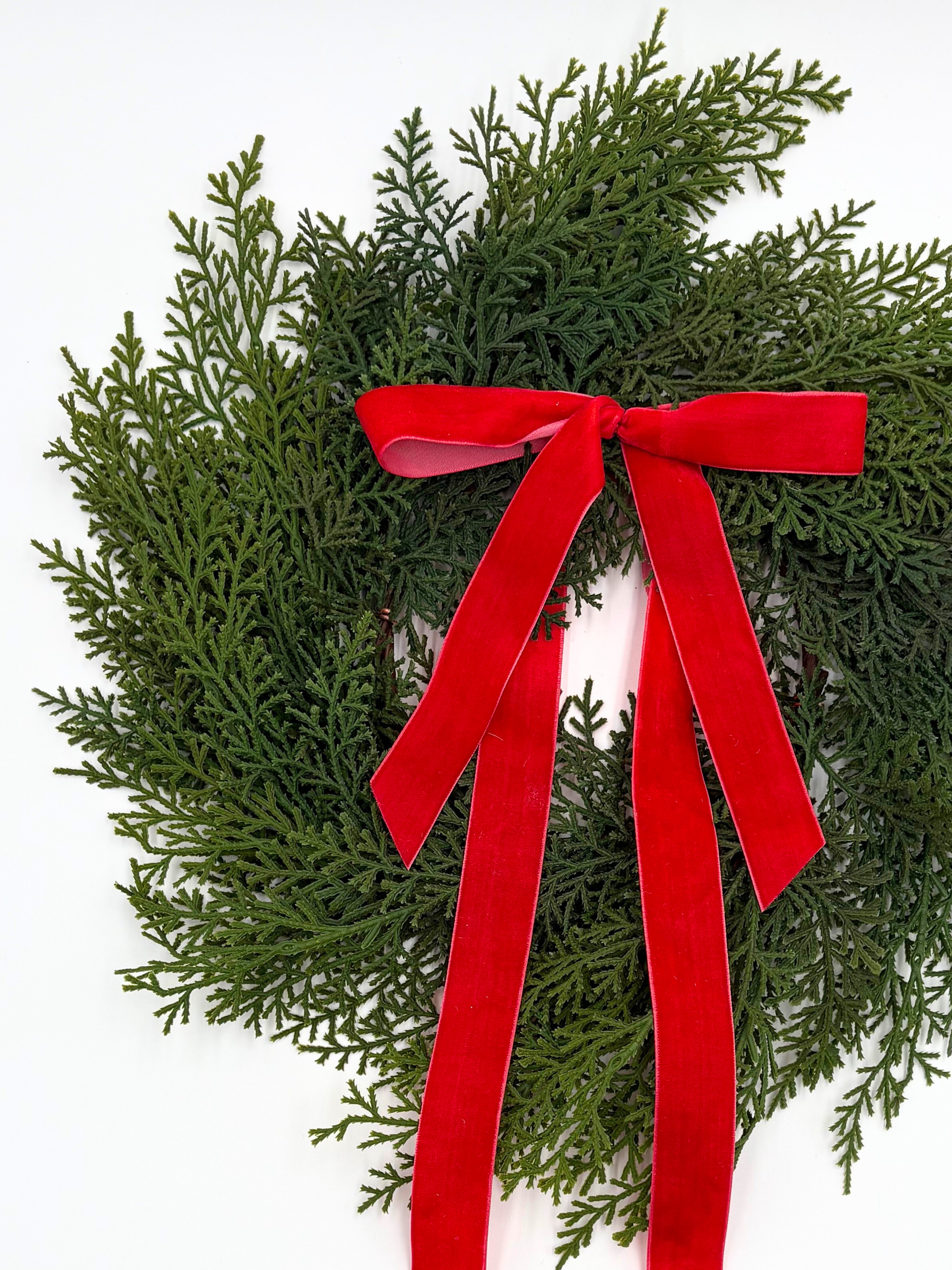 Mini Christmas cabinet wreath made of faux green cedar greenery accented with a long, elegant red velvet ribbon.