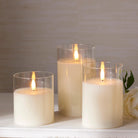 Three ivory LED pillar candles in clear glass holders with realistic flickering flames on a white surface.