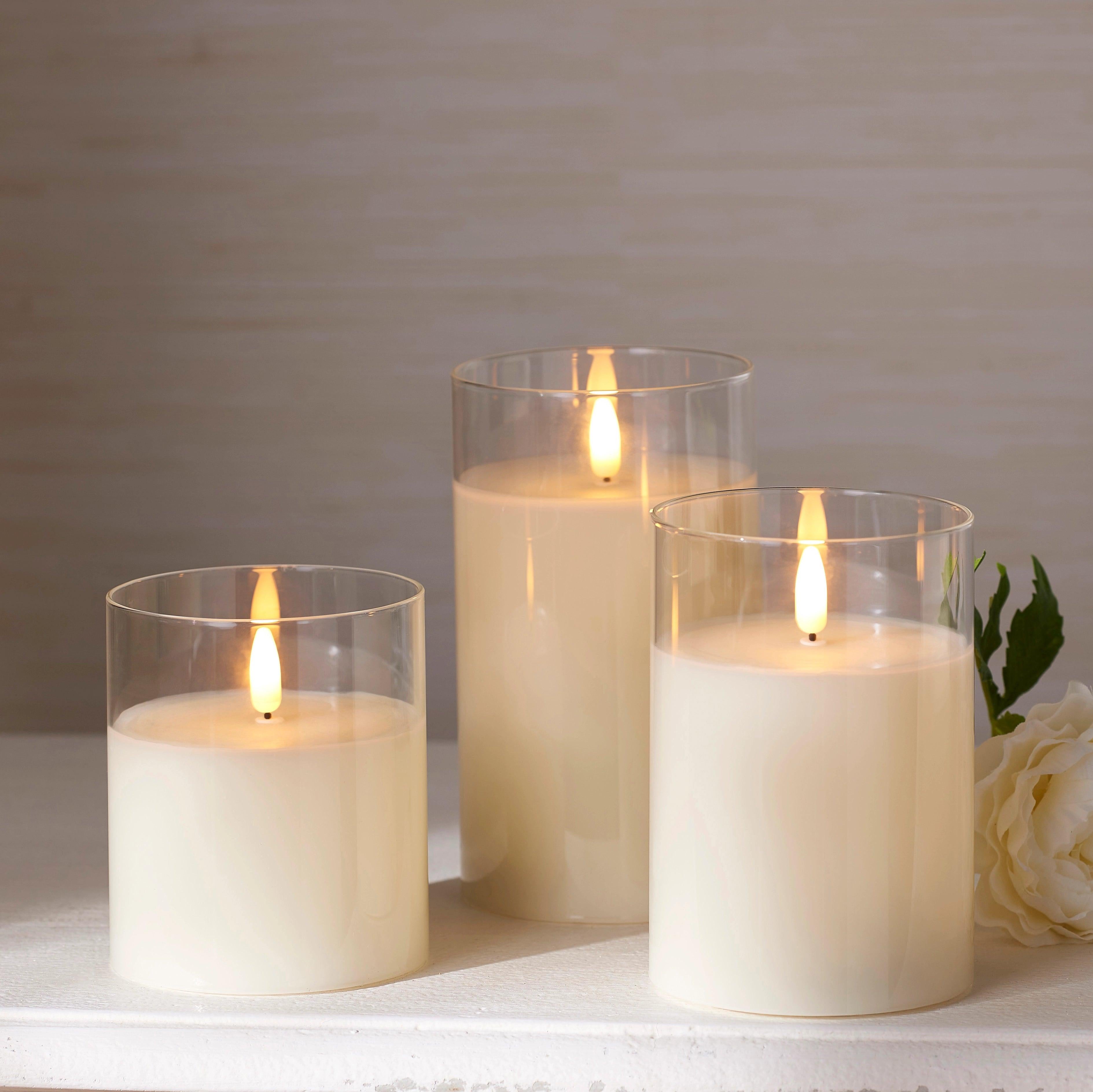 Three ivory LED pillar candles in clear glass holders with realistic flickering flames on a white surface.