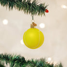 Hand-painted yellow glass tennis ball Christmas ornament with glittery white seams hanging from a pine tree branch.