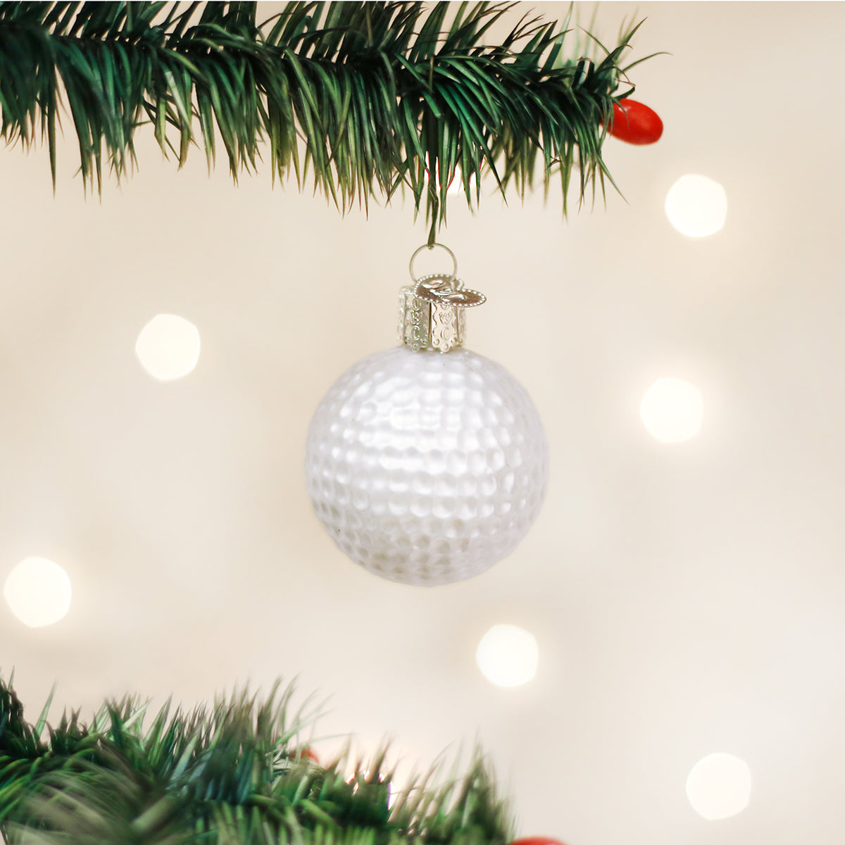 White dimpled golf ball Christmas ornament hanging from a pine branch with a silver cap against a bokeh light background.