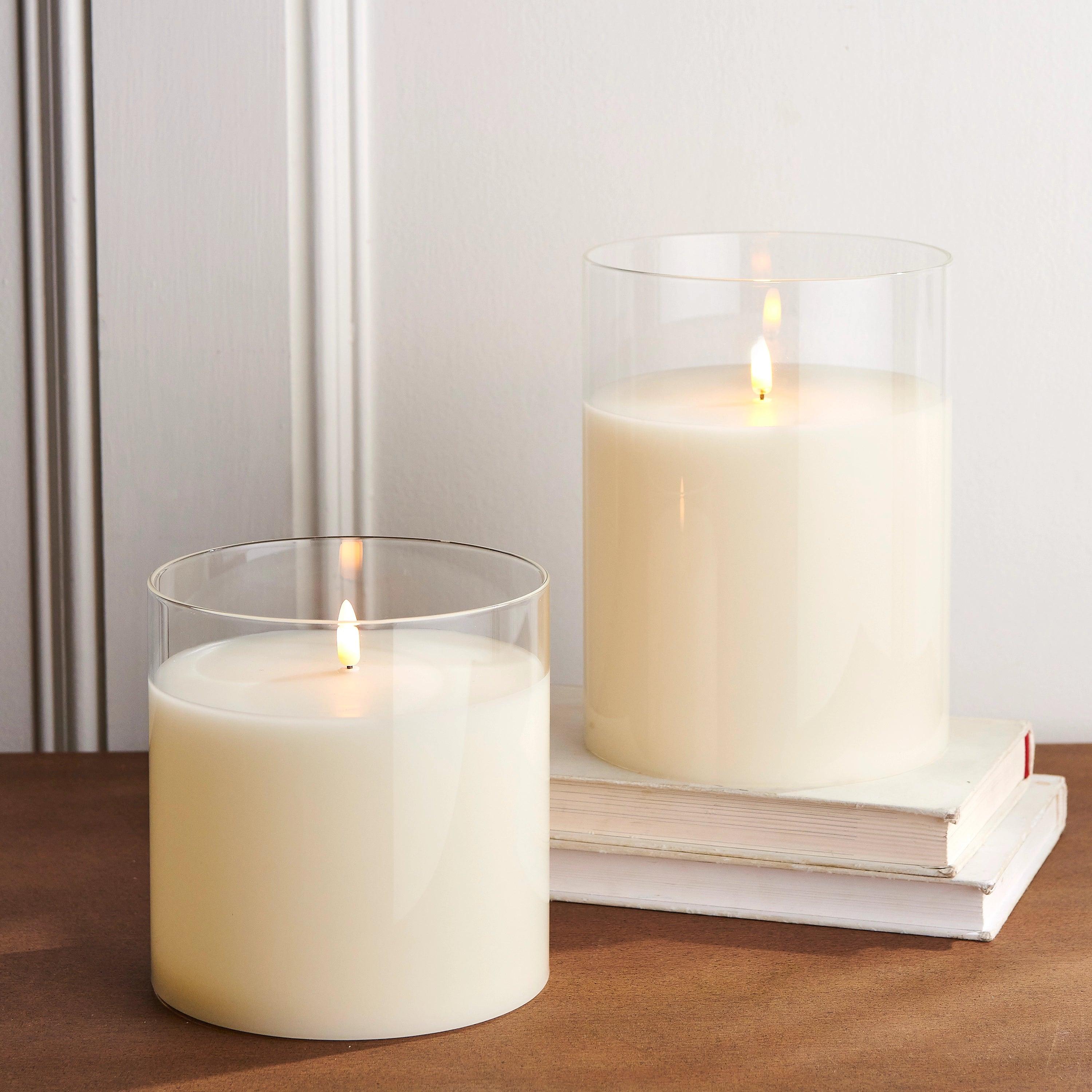 Two ivory LED pillar candles in clear glass holders with realistic flickering flames, set on a wooden surface with books.