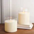 Two ivory LED pillar candles in clear glass holders with realistic flickering flames, set on a wooden surface with books.