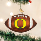 Glass Oregon Ducks football ornament featuring a yellow O logo, white glitter stripes, and a gold heart charm.