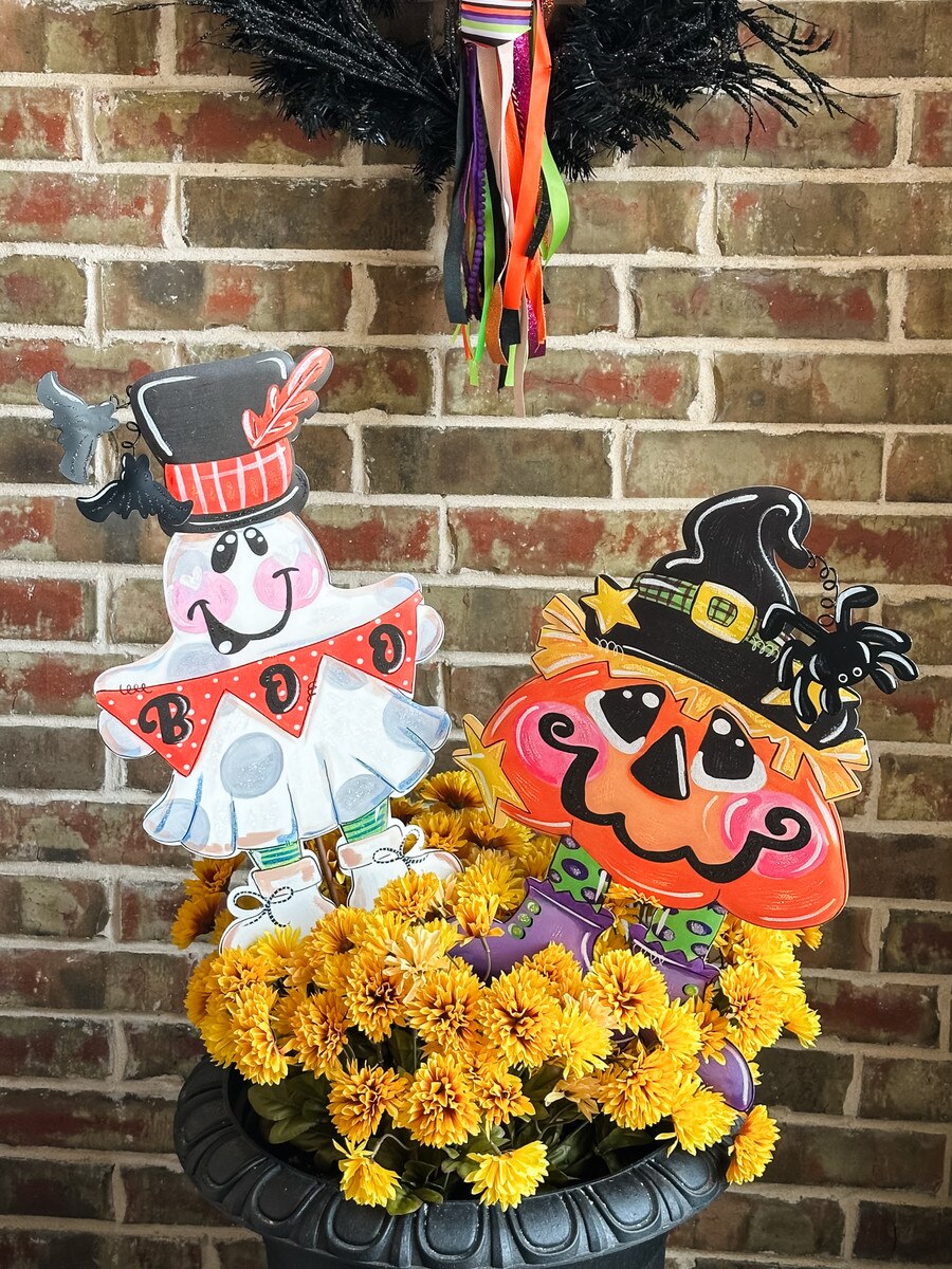 Lori Martin Pumpkin Witch garden stake with a black hat and spider, displayed in a pot of yellow mums against brick.