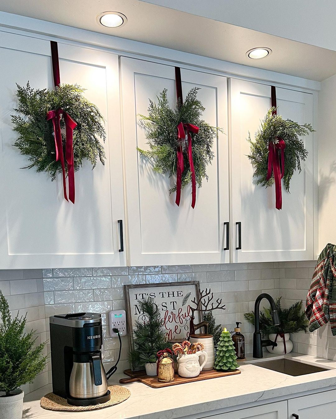 Three mini faux cedar Christmas wreaths with deep red velvet ribbons hanging on white kitchen cabinets.