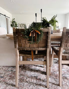 Mini faux cedar wreath with a caramel velvet ribbon hanging on the back of a woven leather dining chair.