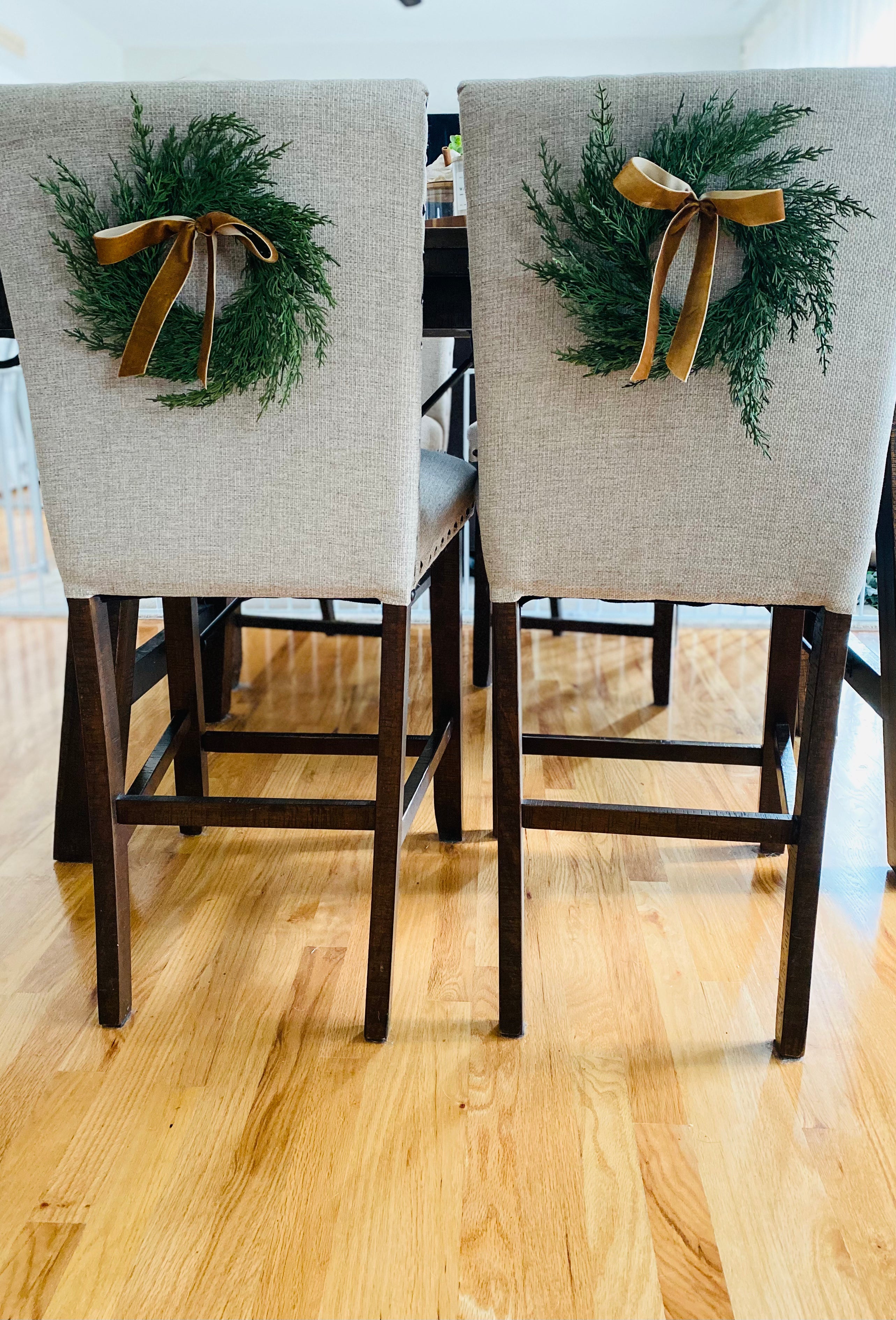 Mini artificial cedar wreaths with elegant gold ribbons hanging on the backs of two upholstered dining chairs.