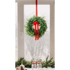 Lifelike Norfolk Pine winter wreath with a red ribbon hanging on a window, featuring lush green needles and holiday decor.