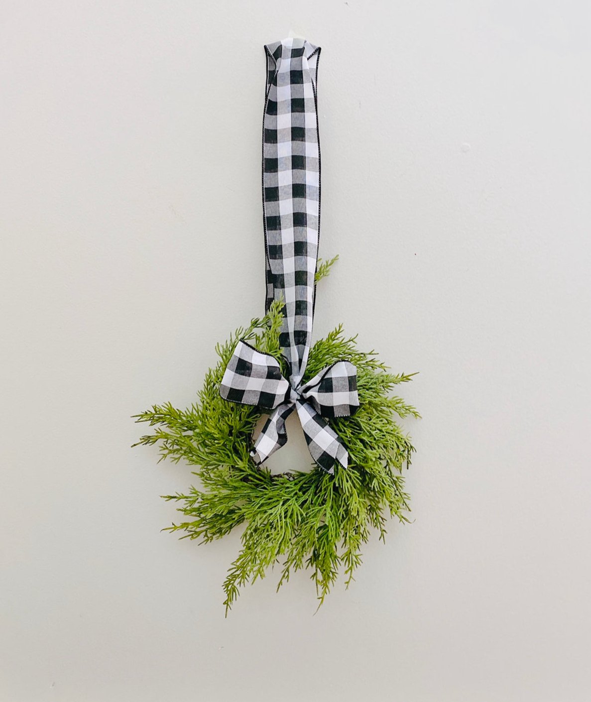 Mini artificial cedar Christmas wreath with a black and white buffalo check ribbon hanging on a plain wall.