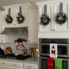 Mini artificial cedar Christmas wreaths with black and white buffalo check ribbons hanging on white kitchen cabinets.