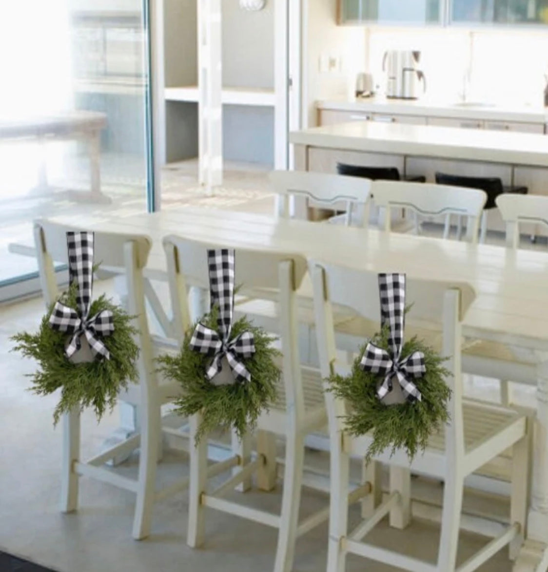 Mini artificial cedar wreaths with black and white buffalo check bows hanging on white dining chairs.
