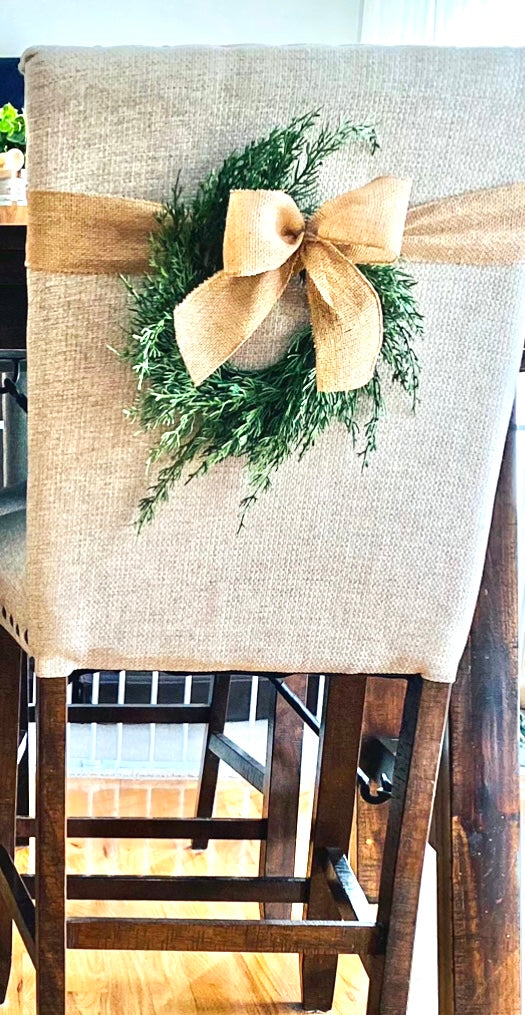 Mini faux cedar Christmas wreath with a rustic burlap bow hanging on a beige upholstered dining chair.