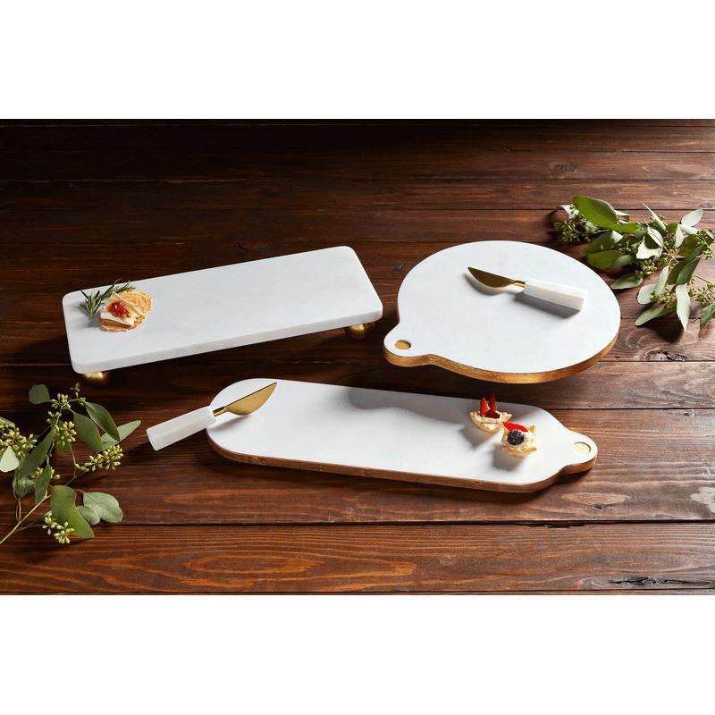 White marble serving boards with gold edges and matching brass spreaders on a dark wood table.